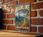 A vintage-style metal sign for the Highline Trail in Montana, featuring a mountain landscape with a trail and flowers.