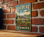 A vintage-style metal sign with the text "Hildene - The Lincoln Family Home" and "The Park is Always Present - Vermont" is displayed on a brick wall.