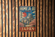 A vintage-style metal sign with Japanese text and an illustration of a Japanese castle or pagoda, mounted on a wooden wall.
