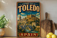 A vintage-style poster of the historic city of Toledo, Spain, featuring a castle and a bridge, is displayed on a wall next to a wooden cutting board and some lemons.