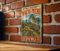 A vintage-style metal sign displaying the name "Historic Sanctuary of Machu Picchu" and the word "Peru" is hanging on a brick wall.