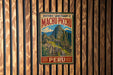 A vintage-style metal sign with the text "HISTORIC SANCTUARY OF MACHU PICCHU PERU" and an illustration of Machu Picchu, a famous Incan citadel, is displayed on a wooden wall.