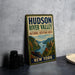 A vintage-style metal sign for the Hudson River Valley National Heritage Area in New York, featuring a scenic illustration of the river valley with a train and buildings.