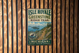 A vintage metal sign for Isle Royale Greenstone Ridge Trail, featuring a scenic mountain landscape with a moose and a trail leading through the forest.