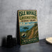 A vintage metal sign for Isle Royale Greenstone Ridge Trail, featuring a scenic illustration of the island and trail, is displayed on a gray countertop next to some black camera lenses.