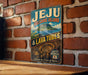 A vintage-style sign for Jeju Volcanic Island, featuring a scenic illustration of the island with lava tubes and a tunnel leading to the volcano.