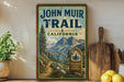 A vintage-style sign for the John Muir Trail in California, featuring a scenic mountain landscape with a trail and a wooden sign.
