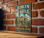 A vintage-style metal sign for "Joslyn Castle" in Nebraska, featuring an illustration of a castle-like building surrounded by trees and a cloudy sky.