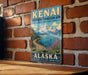A vintage metal sign for the Kenai Mountains Turn Again Arm National Heritage Area in Alaska, featuring a scenic mountain landscape with a river and a small boat.