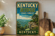 A vintage-style sign for Kentucky Ridge State Forest, featuring a scenic mountain landscape with a cliff and trees.