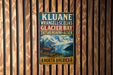 A vintage-style metal sign with text and an illustration of a moose in front of a mountain landscape, mounted on a wooden wall.