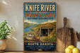A vintage-style sign for Knife River Indian villages, featuring a scenic illustration of the area with a river, mountains, and traditional Native American pottery.