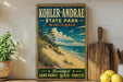 A vintage-style sign for Kohler-Andrae State Park in Wisconsin, featuring a beach scene with sand dunes, trees, and a blue sky with clouds.