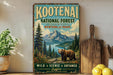 A vintage-style metal sign for Kootenai National Forest, Montana and Idaho, featuring a scenic image of a bear in the foreground with mountains and a lake in the background.