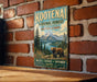 A vintage-style metal sign for Kootenai National Forest in Montana and Idaho, featuring a scenic image of a bear in the foreground with mountains and trees in the background.