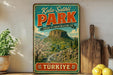 A vintage-style metal sign with a scenic mountain landscape and text in Turkish, placed on a wooden shelf next to a potted plant and a wooden cutting board.