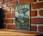 A vintage-style sign for Kuskawa Territorial Park, Yukon, Canada, featuring a mountain landscape with a lake and pine trees.