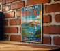 A vintage metal sign with a scenic image of a coastal town, featuring a castle, a sailboat, and flamingos, mounted on a brick wall.