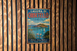 A vintage-style metal sign with the text "LAGUNA DI ORBETELLO ITALIA" and a scenic illustration of a coastal town with a castle, boats, and flamingos.
