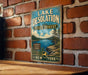 A vintage-style metal sign for Lake Desolation State Forest in New York, featuring a scenic landscape with a lake, mountains, and pine trees.