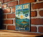 A vintage-style sign for Lake Erie National Marine Sanctuary, featuring a lighthouse, fish, and a scenic background, mounted on a brick wall.
