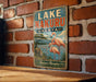 A vintage-style metal sign with the text "LAKE NAKURU KENYA" and an illustration of flamingos and a rhinoceros in a natural setting.