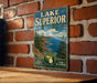 A vintage-style metal sign for Lake Superior State Forest in Michigan, featuring a scenic landscape with pine trees, cliffs, and a body of water.