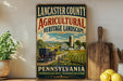 A vintage-style sign for Lancaster County Agricultural Heritage Landscape, featuring a farm scene and a horse-drawn carriage, with the text "LANCASTER COUNTY AGRICULTURAL HERITAGE LANDSCAPE" and "PENNSYLVANIA PRESERVING OUR ROOTS SUSTAINING OUR FUTURE".