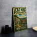 A vintage-style metal sign for the Lead Mine State Forest in Missouri, featuring a scenic trail and camping sign, is displayed on a gray countertop next to some black camera lenses.