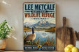 A vintage-style sign for Lee Metcalf National Wildlife Refuge in Montana, featuring a goose standing on a grassy bank with a mountain backdrop.