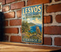 A vintage-style metal sign for LESVOS ISLAND PARK in Greece, featuring an illustration of a scenic landscape with an owl perched on a cliff overlooking the sea.