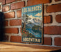 A vintage-style metal sign with a landscape depicting a mountain and lake, mounted on a brick wall.