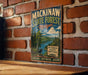 A vintage metal sign for Mackinaw State Forest in Michigan, featuring a scenic illustration of a forest and a moose, is displayed on a brick wall.