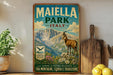 A vintage-style metal sign for Maiella Park in Italy, featuring a mountain landscape with a goat and flowers, placed on a wooden shelf next to a potted plant and a wooden cutting board.