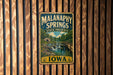 A vintage metal sign for Malanaphy Springs State Preserve in Iowa, featuring a scenic landscape with a waterfall and a wooden bridge.