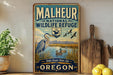 A vintage-style metal sign for the Malheur National Wildlife Refuge in Oregon, featuring an illustration of a heron and ducks in a lake surrounded by mountains.