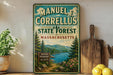 A vintage metal sign for the Manuel Correllus State Forest in Massachusetts, featuring a scenic landscape with pine trees, a lake, and mountains.