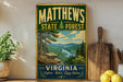 A vintage-style metal sign for Matthews State Forest in Virginia, featuring a scenic mountain landscape with pine trees and a river.