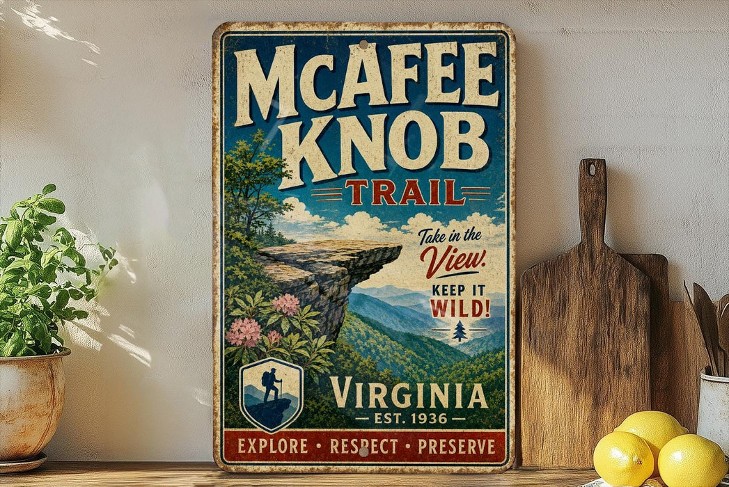A vintage-style metal sign for the Mcafee Knob Trail in Virginia, featuring a scenic mountain landscape and encouraging visitors to "Take in the View! Keep it Wild!" The sign is displayed on a wooden shelf, with a potted plant and a wooden cutting board nearby.
