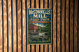 A vintage-style metal sign for McConnells Mill State Park in Pennsylvania, featuring a red brick building and a waterfall, mounted on a wooden wall.