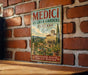 a vintage-style sign for "Medici Villas & Gardens" in Tuscany, Italy, with a brick wall and a wooden table in the background.