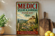 a vintage-style sign advertising "MEDICI VILLAS & GARDENS TUSCANY ITALY" with a scenic illustration of a villa and garden. The sign is displayed on a wall, surrounded by various kitchen utensils and decor items, such as a potted plant, a wooden cutting board, and a bowl of fruit.