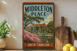 A vintage-style sign for "Middleton Place" in South Carolina, featuring a house surrounded by trees and flowers, is displayed on a shelf next to a wooden cutting board and some potted plants.