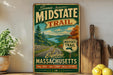A vintage-style metal sign advertising the "Scenic Midstate Trail" in Massachusetts, featuring a scenic mountain landscape and inviting visitors to explore the state.