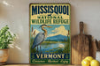 A vintage-style metal sign for the Missoula National Wildlife Refuge in Vermont, featuring an illustration of a heron in a marshy area with mountains in the background.
