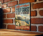 A vintage-style sign for the Mississippi Gulf Coast National Heritage Area, featuring a lighthouse, boats, and a bird, is displayed on a brick wall.