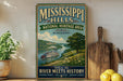 A vintage-style metal sign for the Mississippi Hills National Heritage Area, featuring a scenic illustration of a river and a steamboat, with the text "Where the RIVER MEETS HISTORY" and "NATURAL BEAUTY * RICH HERITAGE * TIMELESS STORIES".