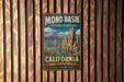 A vintage-style metal sign for the Mono Basin National Scenic Area in California, featuring a scenic landscape with mountains, water, and unique rock formations.