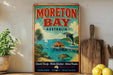 a vintage-style poster of Moreton Bay, Australia, with a tropical beach scene and a dolphin jumping out of the water. The poster is displayed on a shelf next to a wooden cutting board and a potted plant.