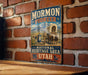 A vintage-style metal sign for the Mormon Pioneer National Heritage Area in Utah, featuring a wagon train and mountain scenery.